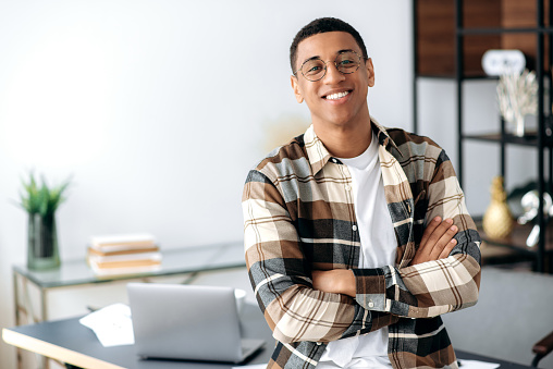 Portrait of a handsome successful confident young mixed race latino man with glasses, stylishly dressed, standing near work desk with arms crossed, looking at camera with friendly smile