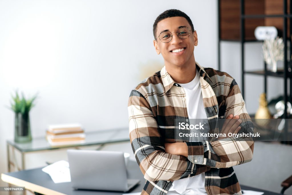Portrait of a handsome successful confident young mixed race latino man with glasses, stylishly dressed, standing near work desk with arms crossed, looking at camera with friendly smile Student Stock Photo