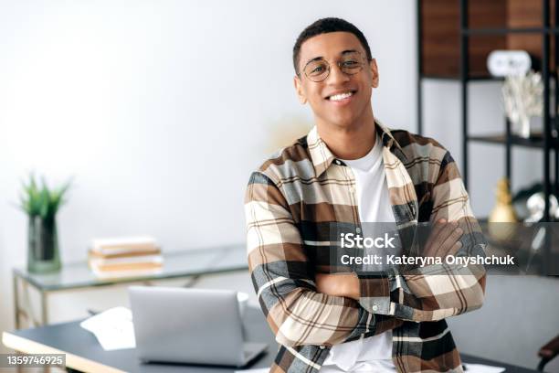 Portrait Of A Handsome Successful Confident Young Mixed Race Latino Man With Glasses Stylishly Dressed Standing Near Work Desk With Arms Crossed Looking At Camera With Friendly Smile Stock Photo - Download Image Now