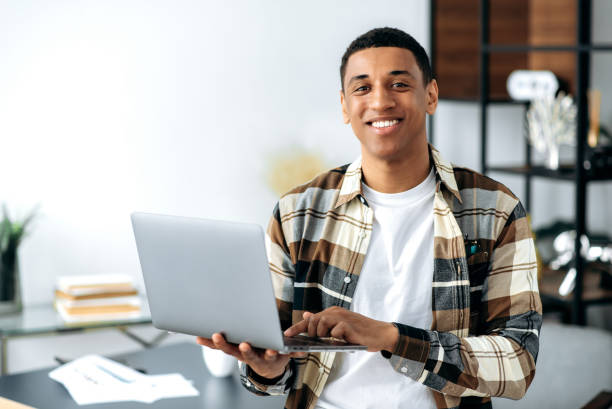 istock Positive handsome charismatic modern hispanic guy, dressed in stylish shirt, office worker, freelancer or student, holding an open laptop, stands near a table, looking at the camera, smiling friendly 1359746927