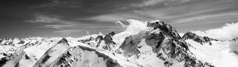Black and white panorama of high snow-capped mountain peaks and beautiful sky with clouds at sun windy day. Caucasus Mountains in winter, region Dombay, mount Dombay-Ulgen.