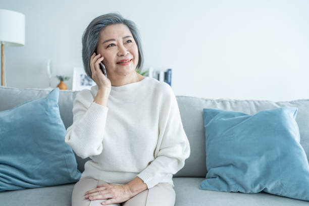 Asian senior elderly woman smile and video call in living room at home. Strong elderly older grandmother feeling happy using mobile cell phone communicate with family enjoy retirement life in house. stock photo