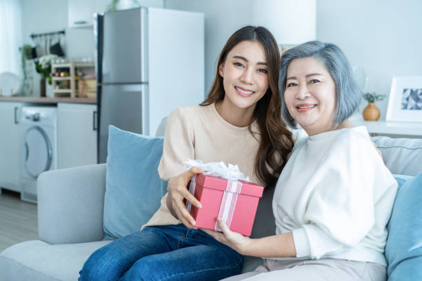 la bella figlia asiatica della donna sorprende sua madre con un regalo di compleanno. giovane ragazza attraente dare la scatola regalo alla vecchia mamma anziana anziana in salotto mentre si siede sul divano in casa. complimenti concetto. - grandmother giving gift child foto e immagini stock