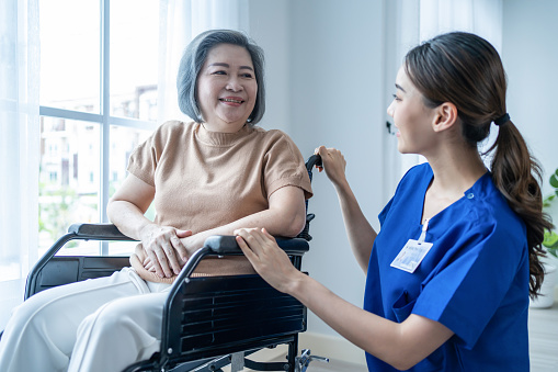 Asian nurse take care senior woman on wheelchair at nursing home care. Attractive happy Caregiver girl doctor take care older elder mature disabled patient female to window in hospital ward to look ou