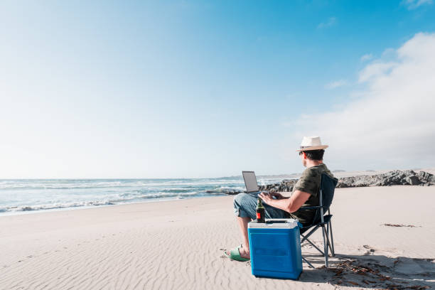 man sitting on camping chair on the beach drinking beer with laptop alone enjoying man sitting on camping chair on the beach drinking beer with laptop alone enjoying digital nomad stock pictures, royalty-free photos & images