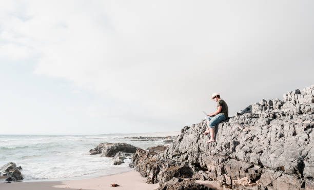 istock man sitting on a rock on the beach with laptop alone doing telecommuting 1359758804