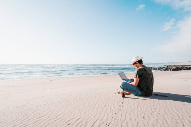 man sitting on the beach drinking beer with laptop alone stock photo