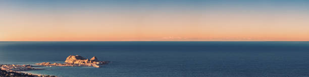 Panoramic view of the red rock of Ile Rousse in Corsica at sunset with the Fench and Italian Alps viewed across the Mediterranean sea in the distance
