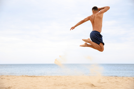 Muscular man jumping on beach, space for text. Body training