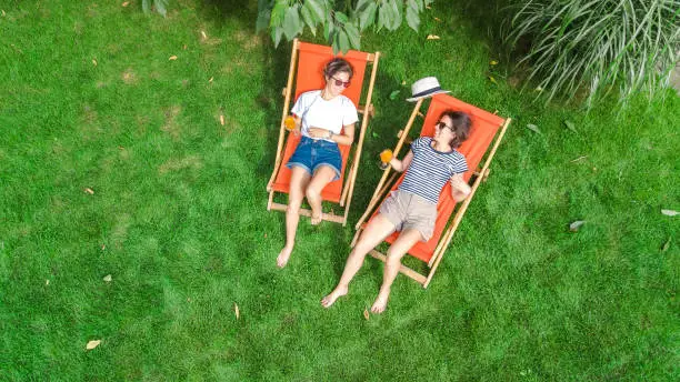 Photo of Young girls relax in summer garden in sunbed deckchairs on grass, women friends have fun outdoors in green park on weekend, aerial top view from above