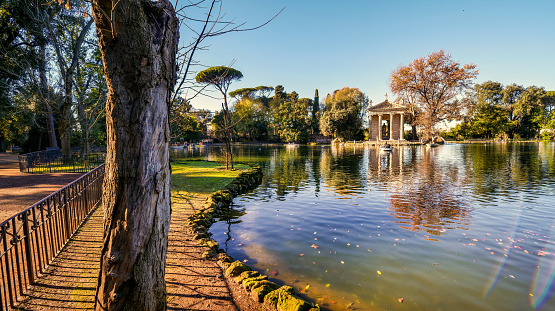 A suggestive and peaceful autumn wide view of Lake Villa Borghese, in the green and historic heart of Rome. In the center, the Ionic-style Temple of Aesculapius, built in 1785 in the gardens of the lake. Considered the green lung of Rome, Villa Borghese is the largest public park in the city with almost 80 hectares of extension, where residents and tourists can spend hours of relaxation and silence among tree-lined avenues, ponds and Italian and English-style gardens surrounded by trees and ancient pines. The area of the Villa Borghese includes the extension of ancient gardens the of the Borghese family since 1580, while the current arrangement of the park was completed in 1903 in neoclassical style. In 1980 the historic center of Rome was declared a World Heritage Site by Unesco. Super wide angle image in high definition and 16:9 format.