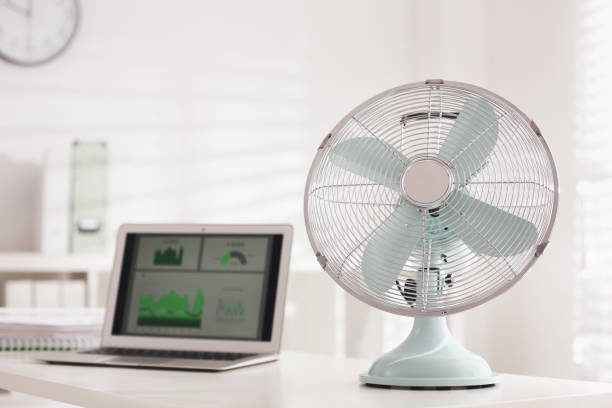 istock Modern electric fan and laptop on table in office 1359815040