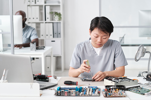 Young Chinese woman in uniform fixing tiny details of circuit board while sitting by desk against her African male colleague