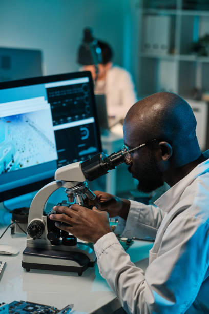 istock Young man of African ethnicity sitting by workplace and looking in microscope 1359815850