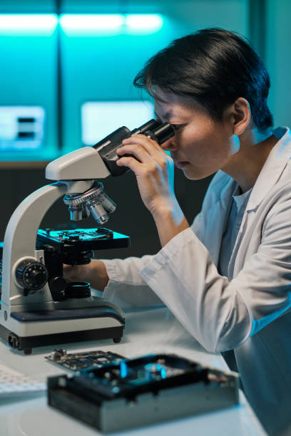 istock Young Chinese woman in whitecoat looking in microscope lens 1359815861