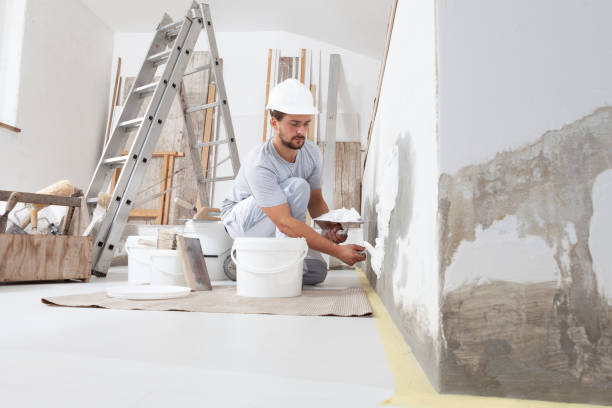 uomo stuccatore operaio edile al lavoro, prende l'intonaco dal secchio e lo mette sulla cazzuola per intonacare il muro, indossa il casco all'interno del cantiere di una casa - cartongesso foto e immagini stock