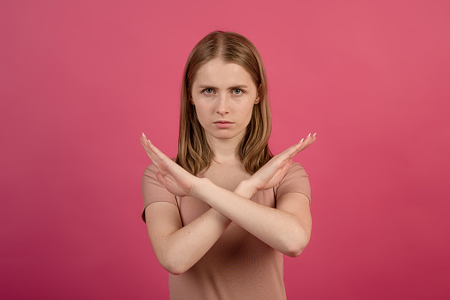 A young and attractive blonde caucasian girl showing a rejection gesture by crossing her arms against a pink studio background.