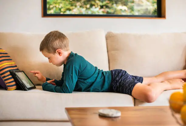 Little boy lying on a living room sofa at home and playing a video game on a digital tablet
