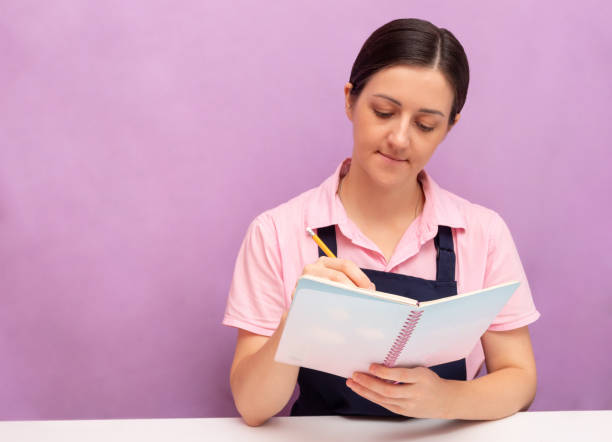 Woman in an apron writes in notebook with pencil. Purple background. Selective focus. Picture for articles about food, confectioners. stock photo