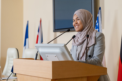 Young successful Muslim businesswoman in hijab standing by tribune while making speech for foreign colleagues at conference