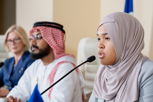Young contemporary female delegate in hijab making speech while sitting against Arabian and American colleagues