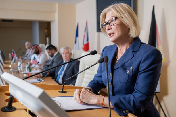 Mature blond delegate in formalwear speaking in microphone Mature blond female delegate in formalwear speaking in microphone while standing by tribune at political forum politician stock pictures, royalty-free photos & images