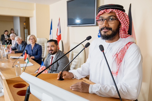Young Arabian speaker looking at camera while standing by tribune with microphone on background of foreign delegates