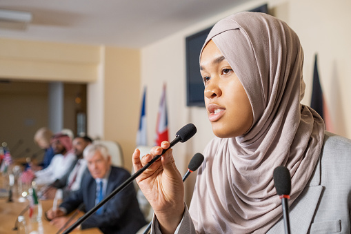 Pretty young Muslim woman in hijab speaking in microphone while standing in front of audience against row of colleagues