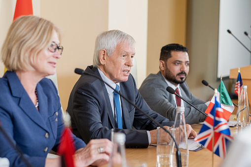 Confident senior politician in formalwear speaking at conference while sitting among his colleagues from foreign countries