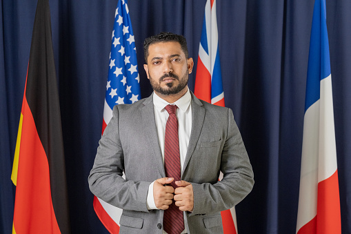 Serious male delegate in elegant suit standing in front of camera against flags of four countries taking part in summit
