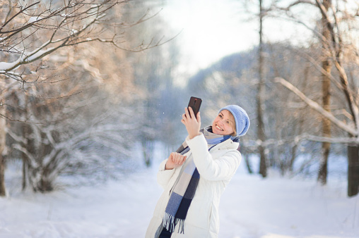 A beautiful blonde woman in a white winter coat and a blue hat walks through a snow-covered park on a winter day and takes pictures on a smartphone and takes a selfie. Winter walks.