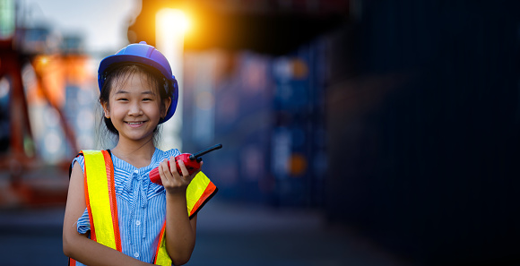 Asian cute girl dressed as an engineer holding walkie-talkie, smiling cheerfully against the backdrop of a large number of shipping containers. Children's ideas, imaginations and goals for the future
