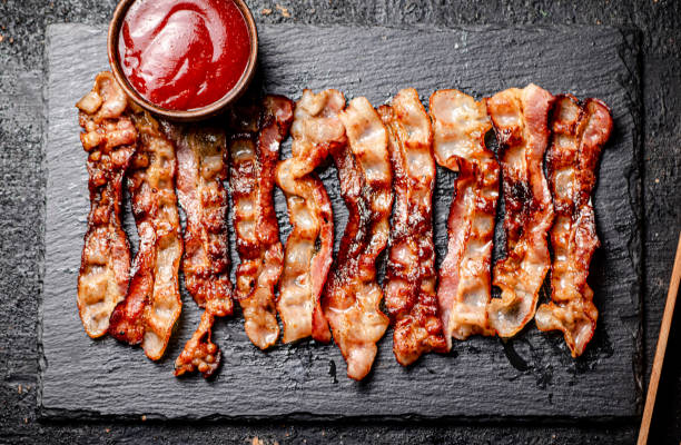 istock Strips of fried bacon on a stone board with tomato sauce. 1359840062