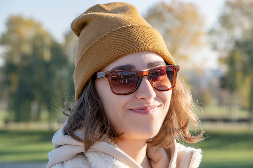 Close-up view of a young female’s portrait. She is at the park for a walk. The weather is sunny and she is happy about that. She is wearing a yellow beanie and sunglasses. She is smiling at the camera.