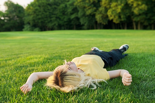 Young blonde woman lying on the grass in a summer park. Teenage female with closed eyes relaxing, resting outdoors. Nature, relaxation, rest, meditation concept
