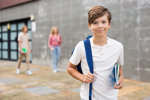 Positive young boy with backpack and copybooks in hand standing at entrance to school building and looking in camera.