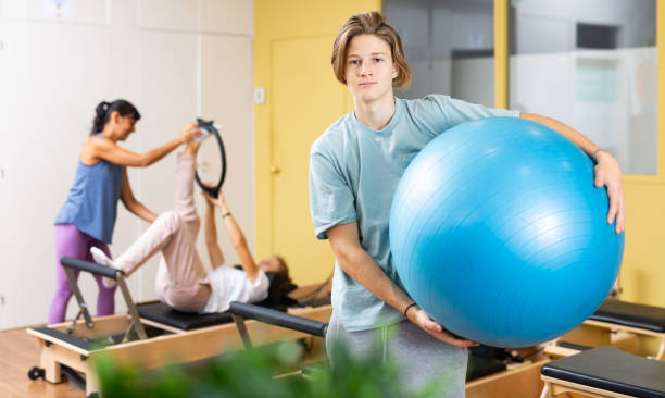 portrait d’un garçon debout avec un ballon de fitness pendant l’entraînement de pilates au gymnase - pilates machine photos et images de collection