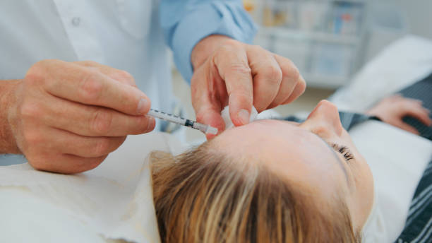 Plastic surgeon doctor making injection botox in woman face under eyebrows on the left side forehead for lifting patient face stock photo