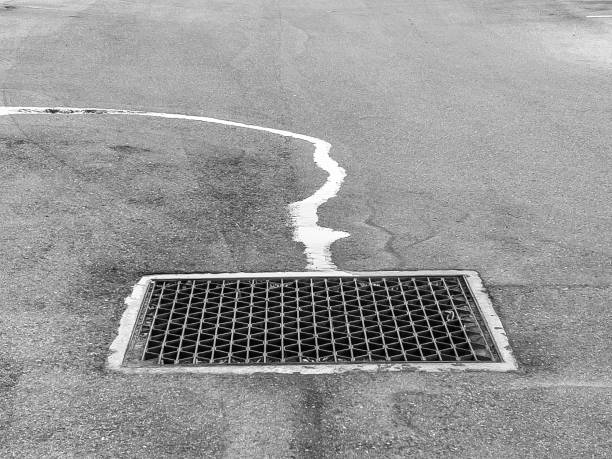 Water runoff reflecting bright sky flows along curve toward steel grate with zigzag pattern over drain in asphalt parking lot, in black and white. For themes of infrastructure and the environment.