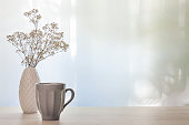 A photo of a desk with a white vase with dried white flowers and a gray cup with coffee.