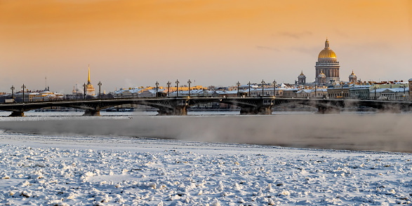 Winter panoramic view of St. Petersburg at sunset, Isaac cathedral and Blagoveshenskiy bridge on background, steam over frozen Neva river, sky of orange color