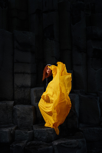 Stately redhead beautiful woman flying her yellow long skirt with basalt columns in the back, Iceland