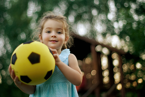 Beautiful little girl , outside in the park holding a yellow soccer footbsll in her hands , smiling and facing camera