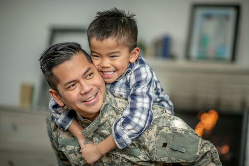 A young boy wraps his arms around his military fathers neck as he Piggy Backs him. The boy is dressed casually and the Father is in military attire as the two embrace tightly after a long deployment overseas.  The boy has a big smile on his face as he is happy to see his father.