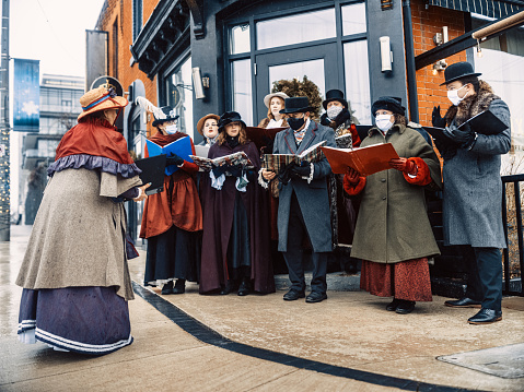 Choir of carol singers performing on the street, during Christmas Holiday. Dressed in historic costumes from 19. century. Some performers wearing face protection masks during performance. Exterior of urban city street with snow falling.