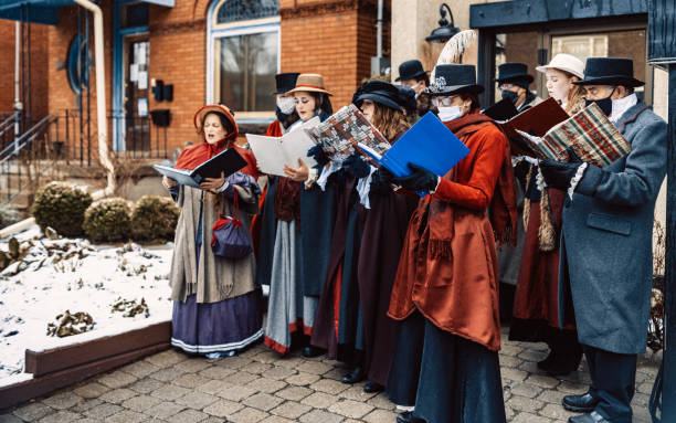 Choir of carol singers performing on the street Choir of carol singers performing on the street, during Christmas Holiday. Dressed in historic costumes from 19. century. Some wearing face protection masks during performance. Exterior of urban city street with snow falling. traditional song stock pictures, royalty-free photos & images