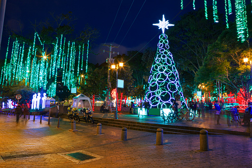 Bogota, Colombia - A Corner of the public Usaquen Town Square, in Bogotá the capital city of Colombia in South America, has been decorated and lit up in LEDs for Christmas celebration. The illumination draws a lot of crowds both local and tourists. Shadows and silhouttes of people walking around the square, can be seen in the photograph. A long exposure creates movement in the photograph and motion blur.  The movement of hanging lights create streaks instead of single bulbs. Horizontal format. Copy space.