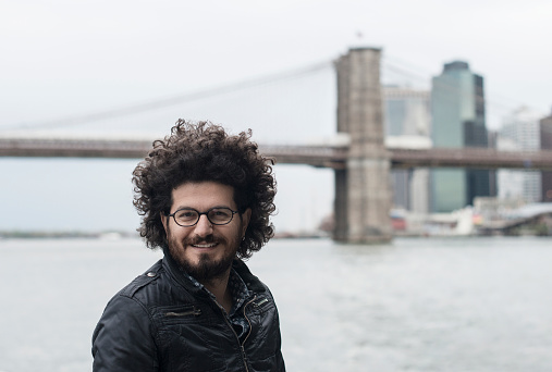 Portrait of a curly haired young man in New York City. Brooklyn Bridge seen on the background