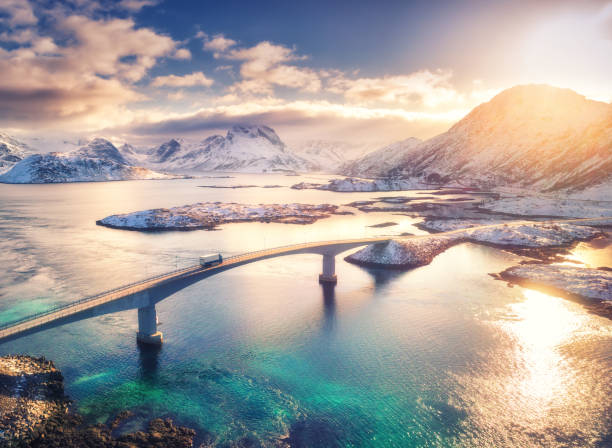 ノルウェーのロフォーテン諸島の橋、海、雪山の空中写真。冬の日没時にフレバン橋。青い水、雪の中の岩、道路、雲のある空の風景。ドローンからのトップビュー - road beach sky cloud ストックフォトと画像