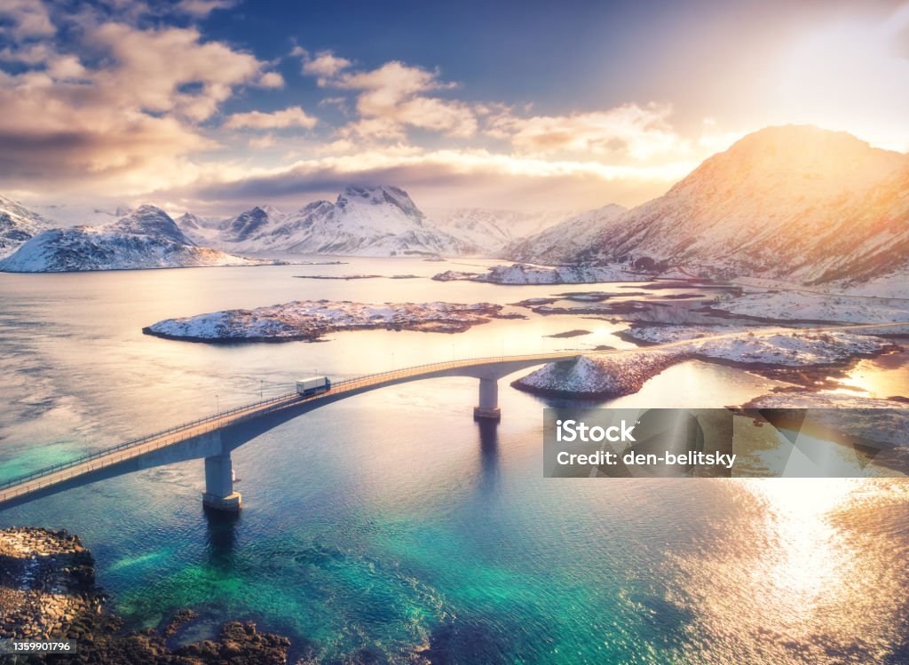 Aerial view of bridge, sea and snowy mountains in Lofoten Islands, Norway. Fredvang bridges at sunset in winter. Landscape with blue water, rocks in snow, road and sky with clouds. Top view from drone Norway Stock Photo